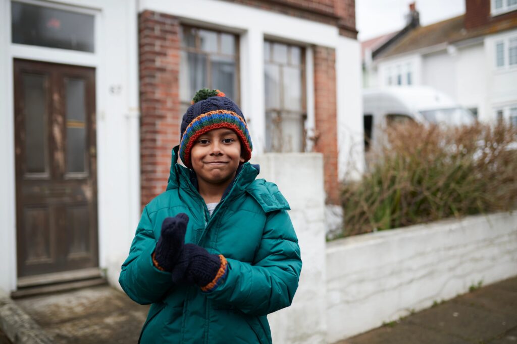 Image of young boy aged around 10, wearing a coat and hat and standing outside a house. He is smiling at the camera whilst putting on one of his gloves.